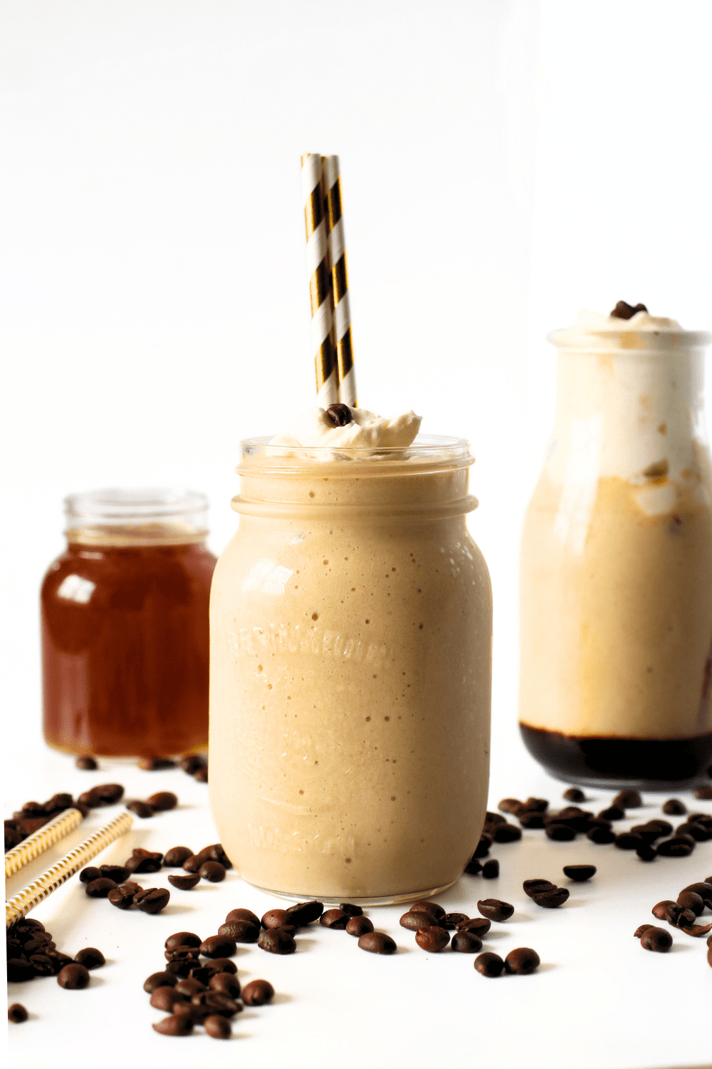 a mason jar with 2 straws filled homemade keto iced cappuccino with homemade sugar-free vanilla syrup in a jar in the background with a second cup of keto iced coffee with sugar-free whipped cream.  Espresso beans are scattered throughout