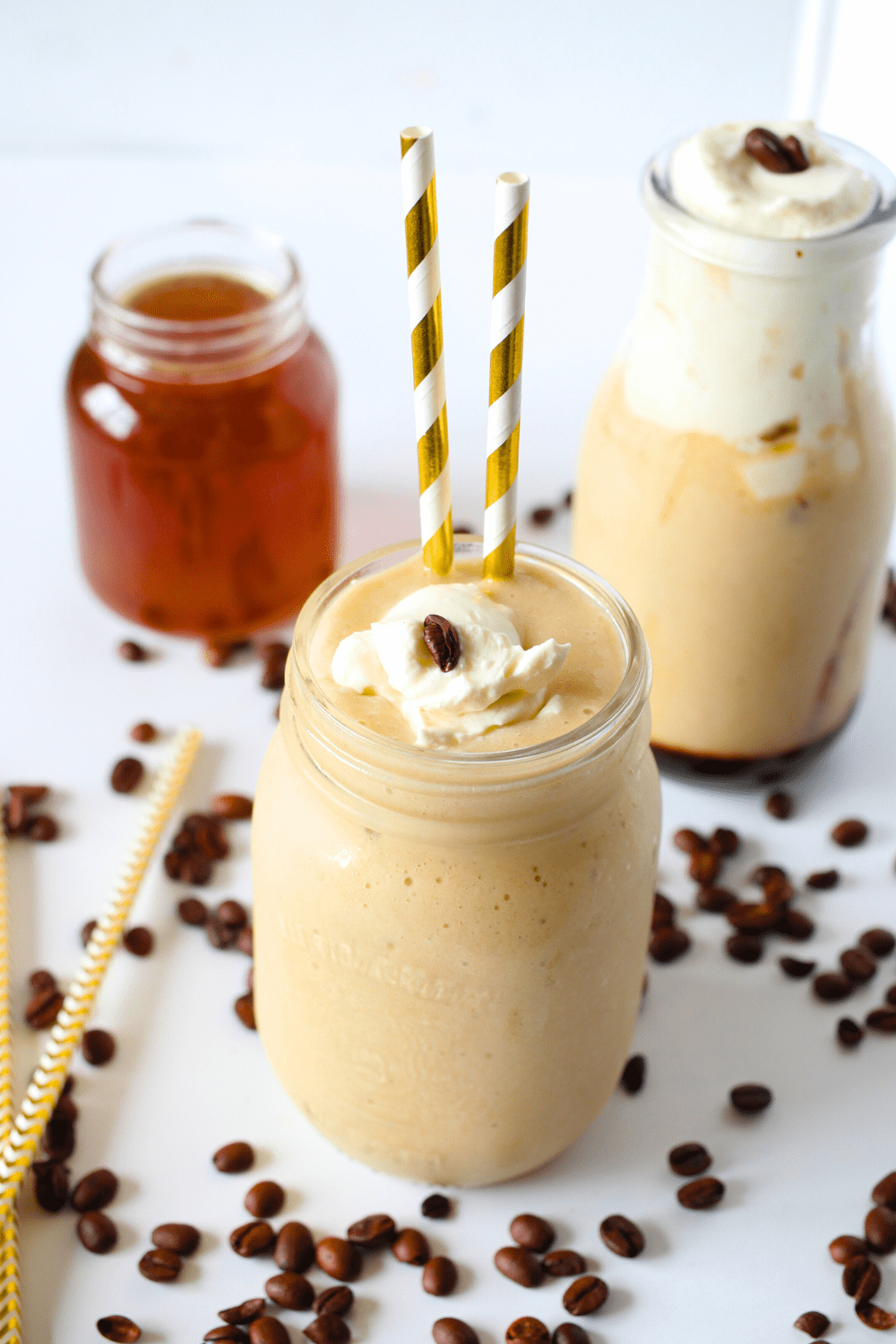 a glass filled with homemade keto iced coffee and two straws with a little whipped cream.  In the background is a second coffee along with sugar-free vanilla coffee syrup and espresso beans scattered throughout
