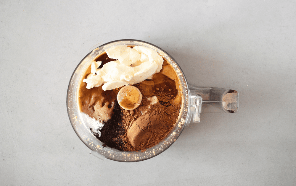 top view of food processor bowl with hazelnut butter, palm shortening, cocoa powder, espresso powder and sweetener added