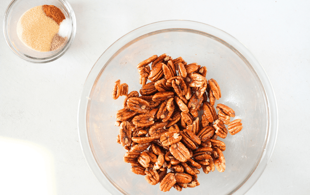 top view of a bowl of pecans tossed in sugar-free maple syrup.  Sugar-free sweetener, salt, chili spice and cinnamon in a small bowl to the side
