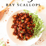 top view of air fryer spicy bay scallops in a bowl with a wooden spoon, herbs and spices scattered alongside the keto scallops