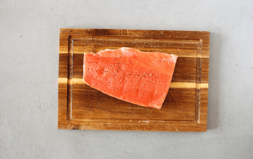 top view of a piece of raw salmon sprinkled with sat and pepper on a cutting board