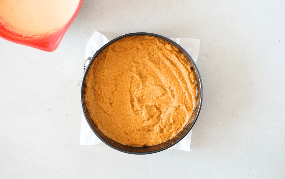 top view of a springform pan lined with parchment paper and keto pumpkin cake in the bottom of the pan.  A separte bowl filled with sugar-free cheesecake batter is to the left.