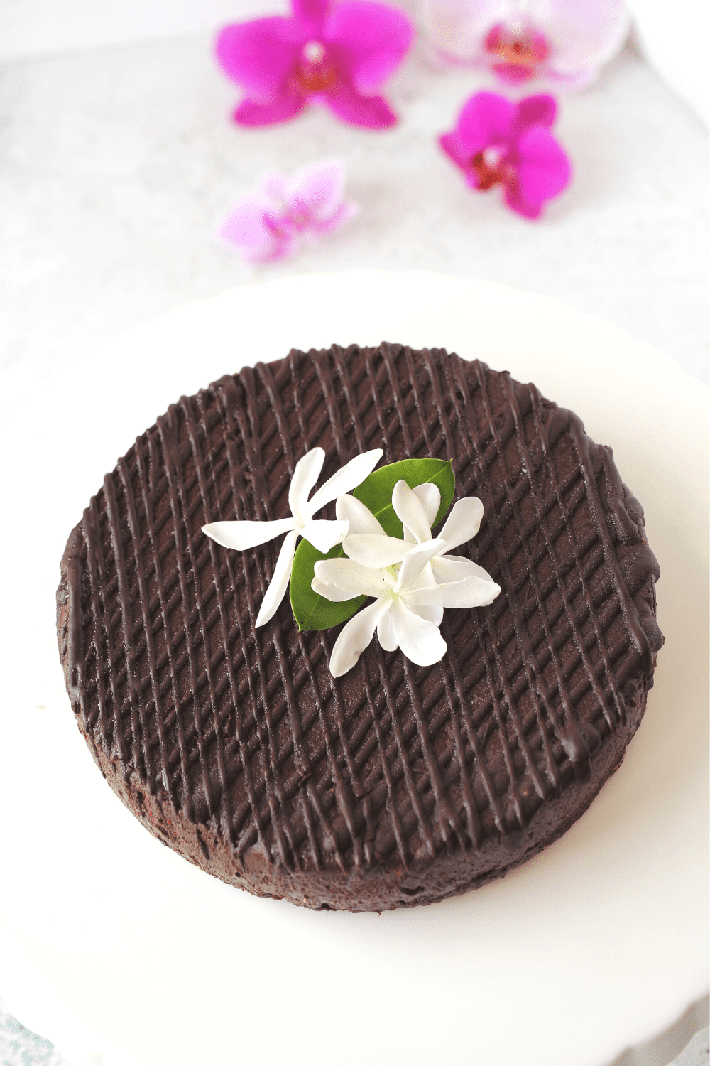 a sugar-free flourless cake on a cake stand wtih chocolate sugar-free ganache drizzled over the gluten-free chocolate cake.  Flowers adorn the cake as decorations