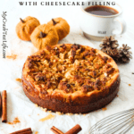a angled view of a keto pumpkin cream cheese crumb cake with a cup of coffee, cinnamon sticks, pumpkins and a whisk lying around the keto pumpkin cake
