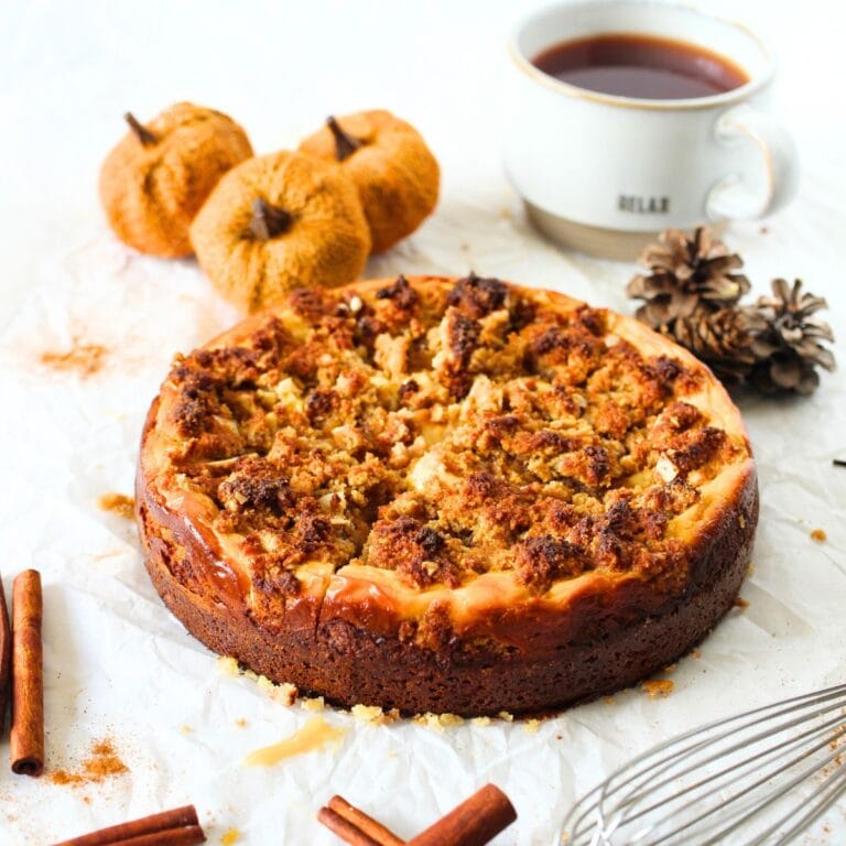 a angled view of a keto pumpkin cream cheese crumb cake with a cup of coffee, cinnamon sticks, pumpkins and a whisk lying around the keto pumpkin cake