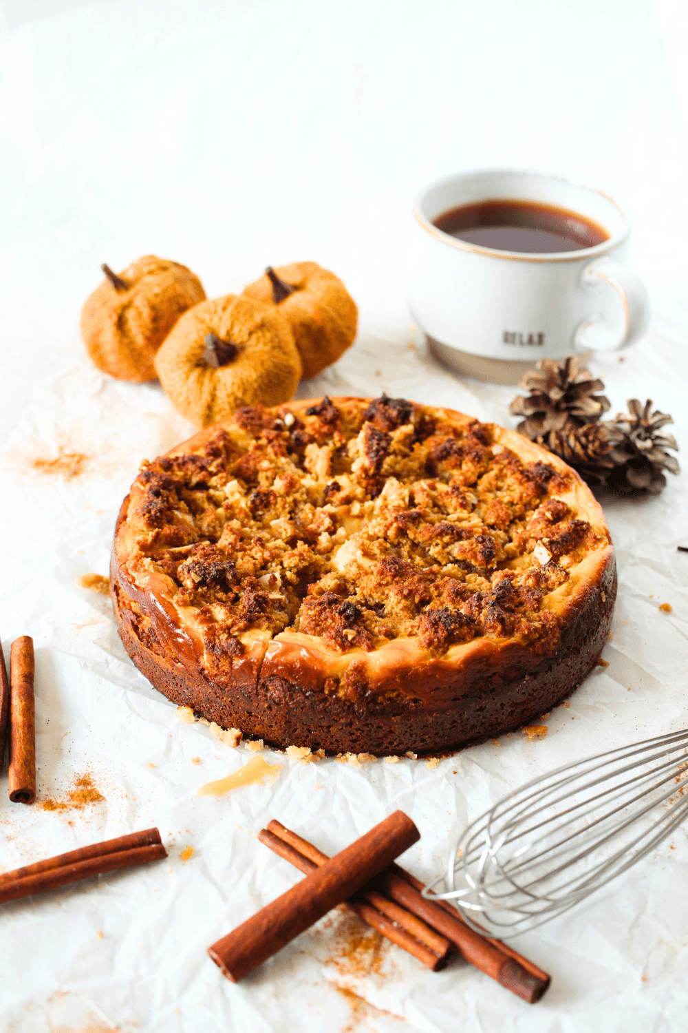 a angled view of a keto pumpkin cream cheese crumb cake with a cup of coffee, cinnamon sticks, pumpkins and a whisk lying around the keto pumpkin cake