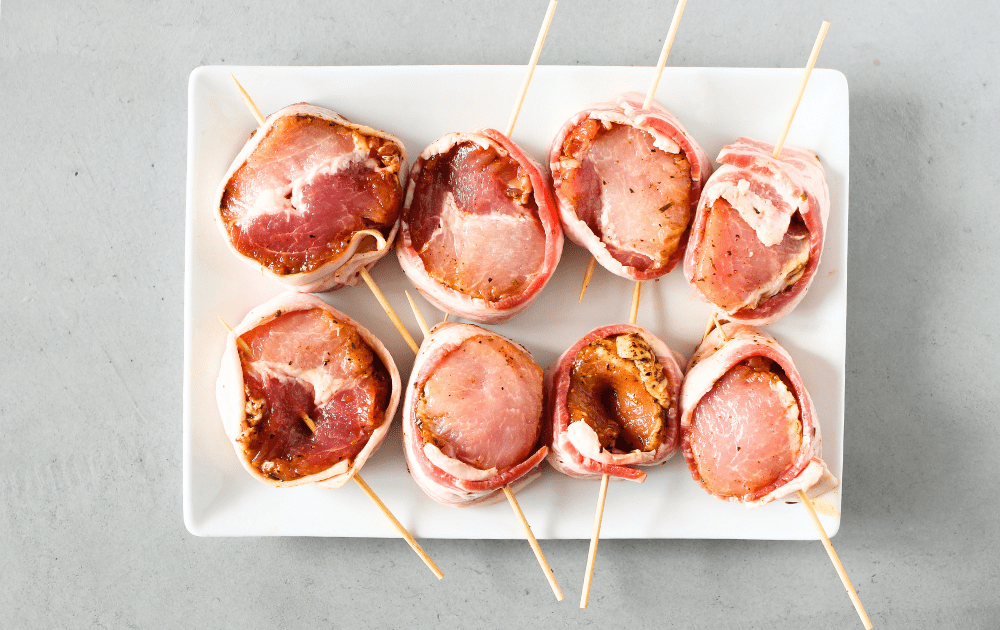 top view of a white place with sections of keto bacon-wrapped pork tenderloins that have been skewered to hold the bacon around the pork tenderloin