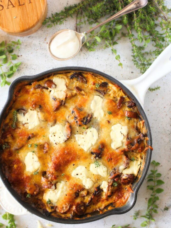 top view of the BEST keto Frittata with sausage, mushrooms and cheese. The keto frittata is dolloped with sour cream. Beside the frittata are fresh herbs, a whisk, raw mushrooms and a spoonful of sour cream