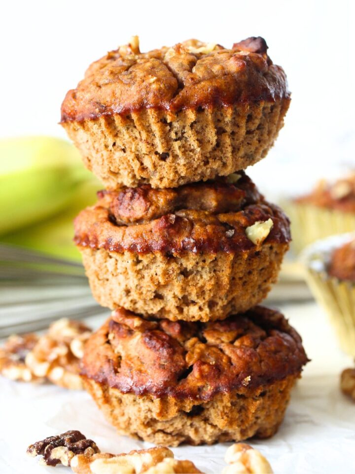 3 oat fiber banana muffins stacked on top of each other with a banana in the background and walnuts scattered throughout