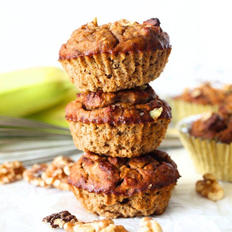 3 oat fiber banana muffins stacked on top of each other with a banana in the background and walnuts scattered throughout