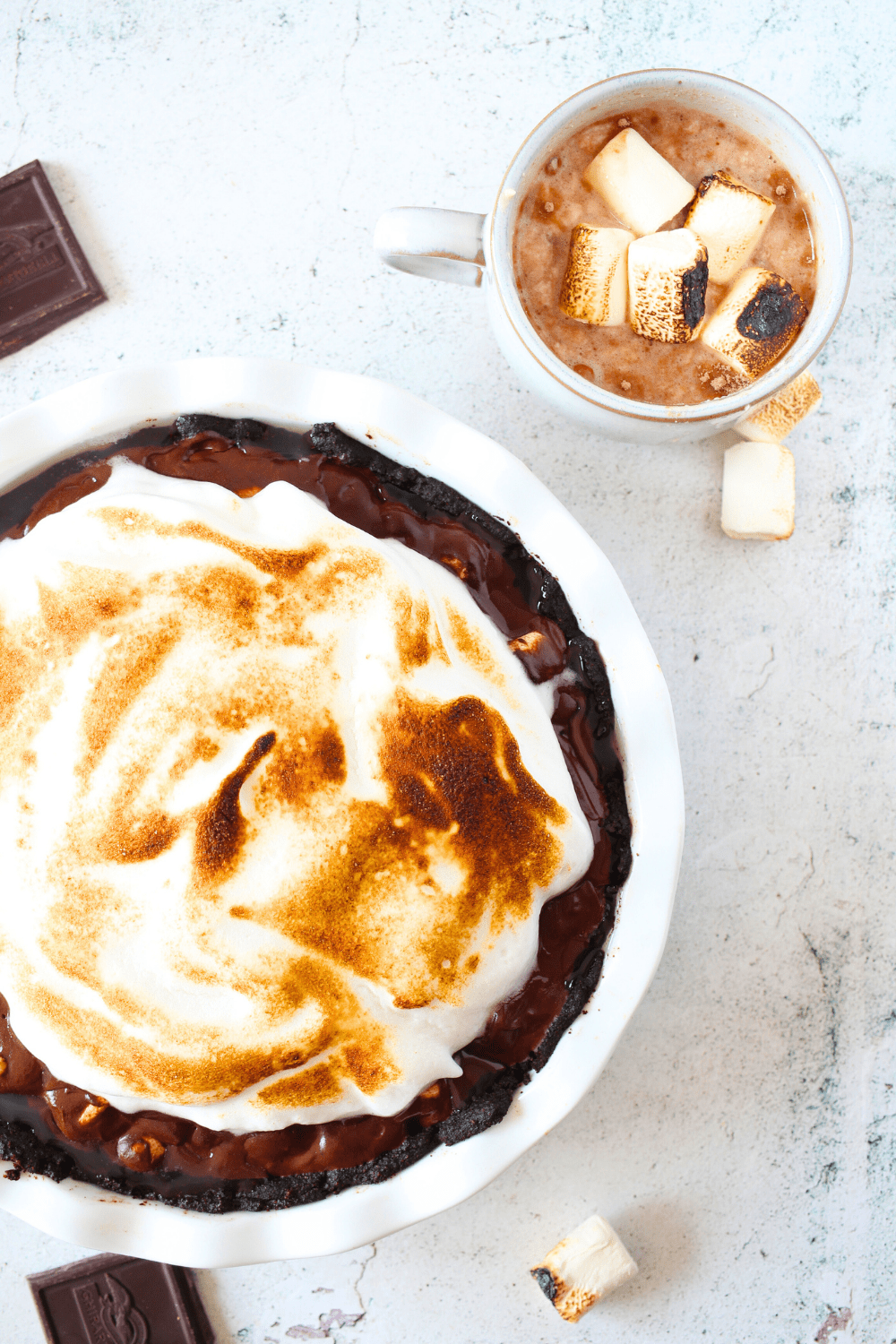 top view of a keto chocolate pie with sugar-free marshmallow frosting, and a cup of hot cocoa with chunks of chocolate scattered around the keto chocolate pie.