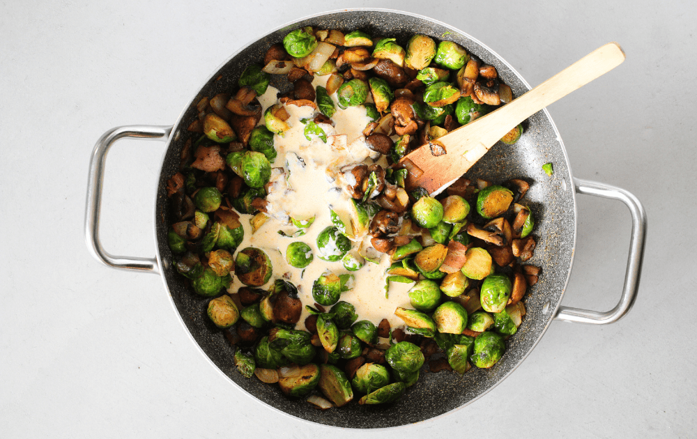 top view of a skillet with roasted Brussels sprouts, mushrooms, onions, bacon and heavy cream being added to the skillet