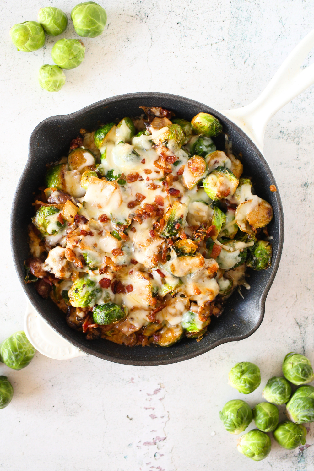top view of a skillet filled with keto cheesy Brussels sprouts casserole with bacon, mushroom and onion. Brussels sprouts scattered around the easy keto side dish
