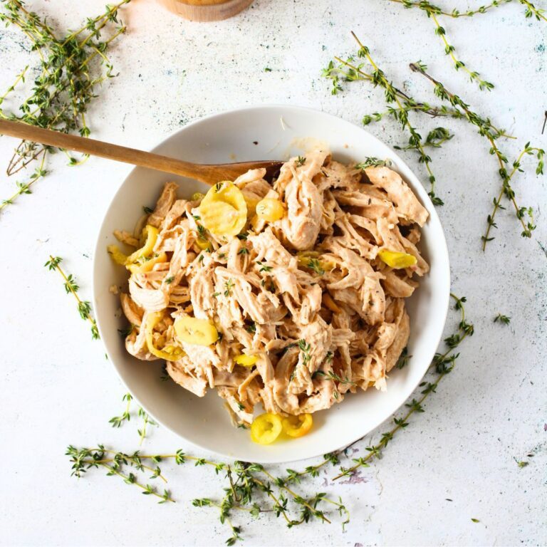 top view of a white bowl filled with keto pulled alabama chicken that is made in an instant pot or slow cooker. There are herbs, banana peppers and a salt container scattered around