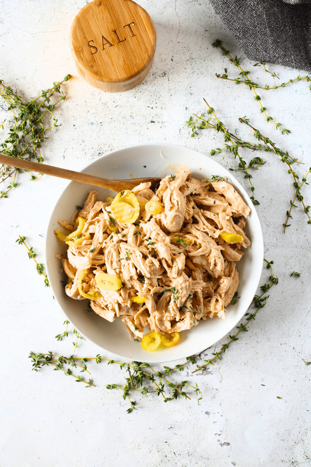 top view of a white bowl filled with keto pulled alabama chicken that is made in an instant pot or slow cooker.  There are herbs, banana peppers and a salt container scattered around