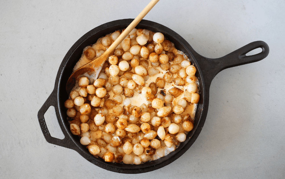 a skillet with browned pearl onions and heavy cream being stirred with a wooden spoon