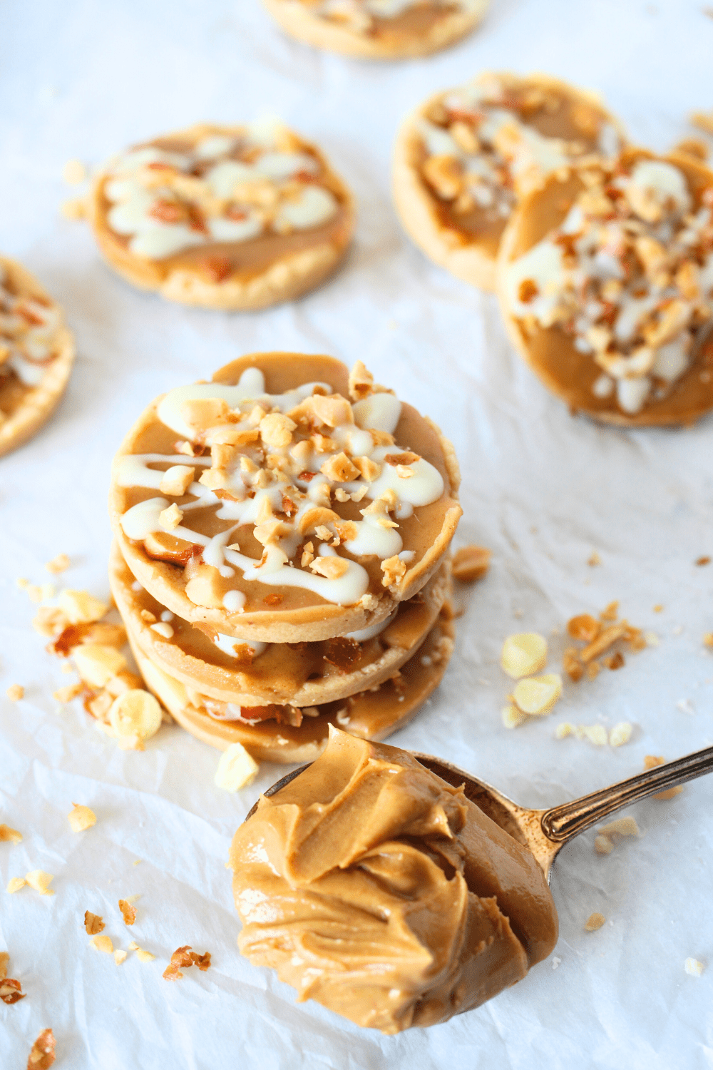 front view of 3 keto white chocoalte peanut caramel cup cookies stacked on top of each other with a spoon of peanut butter and nuts scattered about.