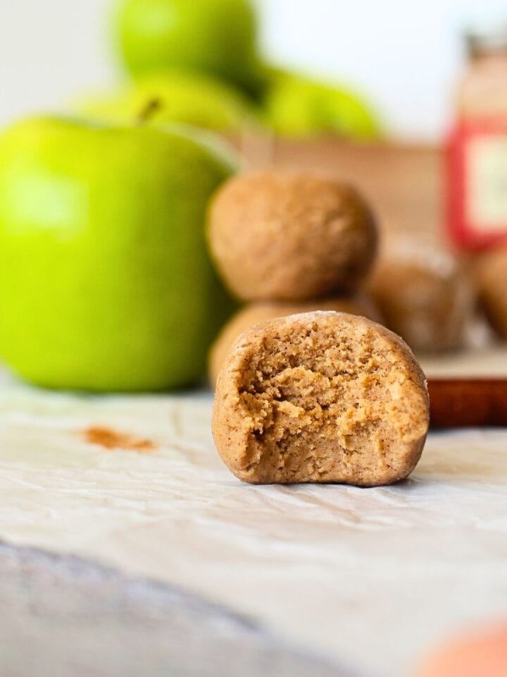 front view of the inside of a apple pie protein bites with green apples in the background as well as apple pie mix and cinnamon sticks