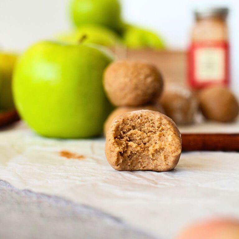front view of the inside of a apple pie protein bites with green apples in the background as well as apple pie mix and cinnamon sticks
