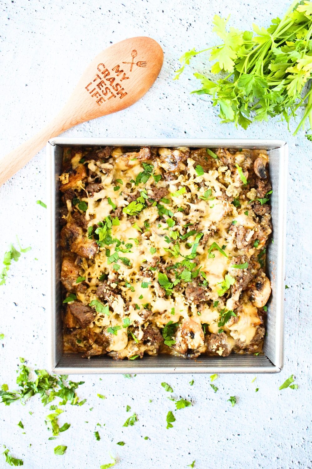 top view of a keto casserole made with sausage and mushrooms.  A wooden spoon and herbs lie alongside the keto casserole