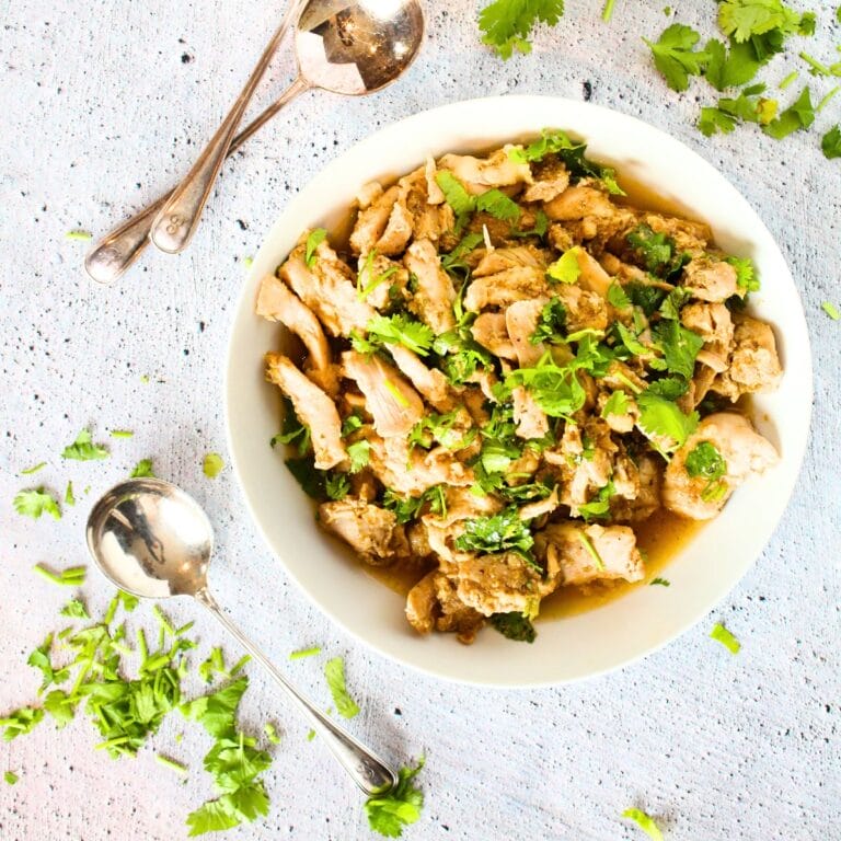 top view of high protein slow cooker sweet cilantro chicken in a bowl with large spoons and fresh cilantro surrounding bowl.