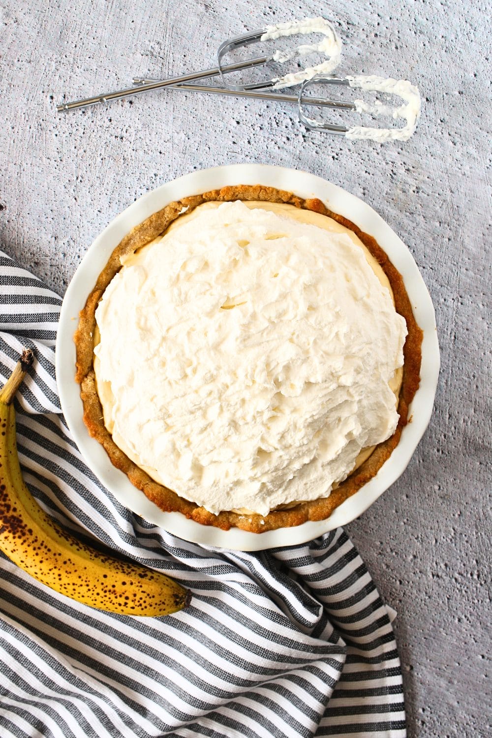 top view of keto banana cream pie in a sugar-free vanilla cookie pie crust with a banana and beaters to the side.