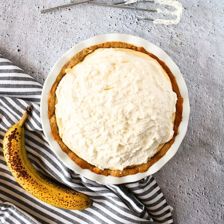 top view of keto banana cream pie in a sugar-free vanilla cookie pie crust with a banana and beaters to the side.
